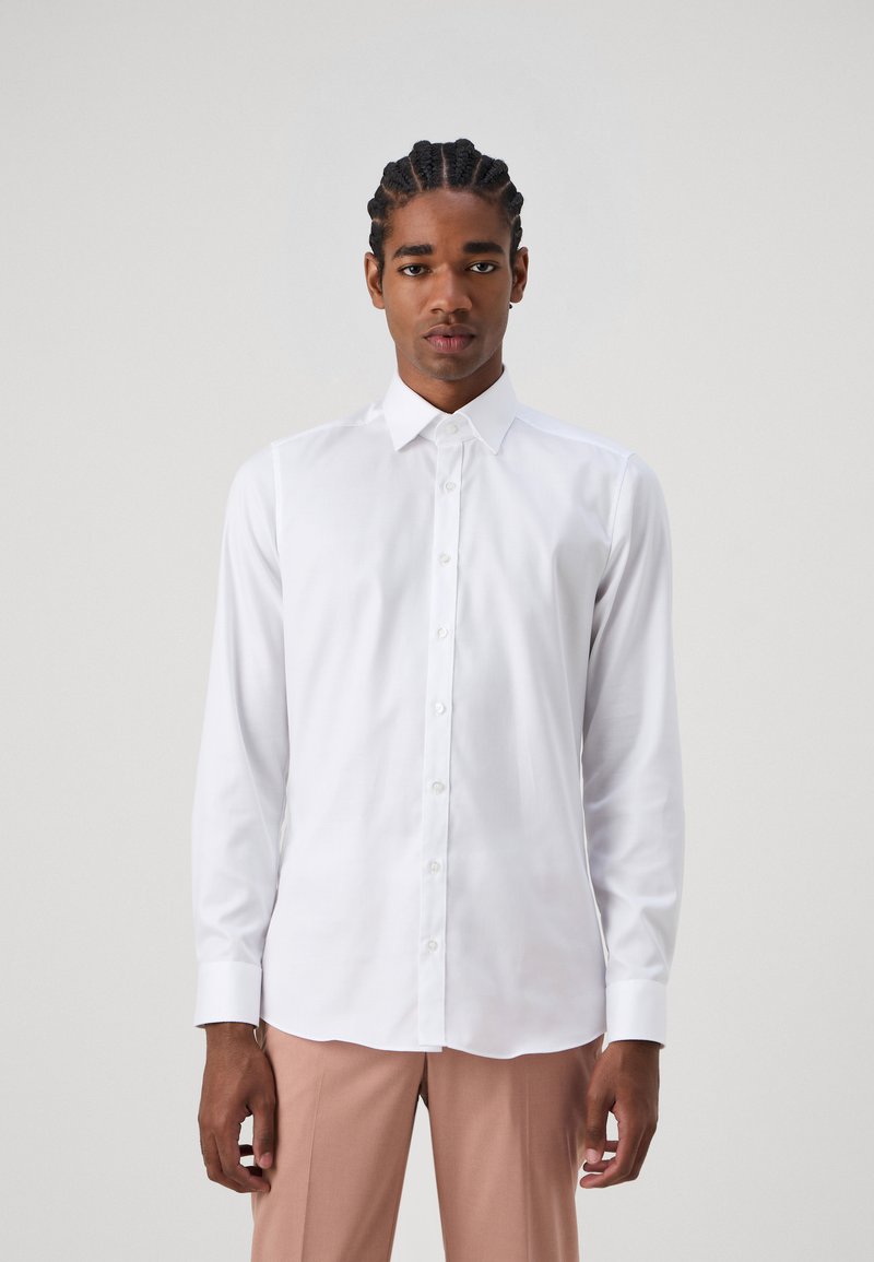 Chemise blanche à manches longues avec un col classique et des boutons sur le devant. Tissu lisse, coupe ajustée et design épuré. Associée à un pantalon léger rose.