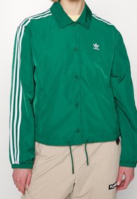 Chaqueta ligera verde con cuello, botones a presión, mangas de rayas blancas, puños elásticos y dobladillo ajustable con cordón. Presenta el logo de Adidas.