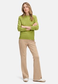 Zöld pulóver bordás mandzsettával, bézs velúrból készült bő szárú nadrággal és fehér lábbelivel párosítva. A modell karba tett kézzel áll.