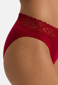 Rode thong met kanten rand, voorzien van een zachte stoffen body en een elastische tailleband. De kant heeft een bloemenpatroon langs de bovenrand.