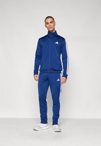 Modra trenirka iz gladkega materiala, ki ima bele črte ob straneh, z zip jopico in Adidasovo logo na prsih in hlačah.