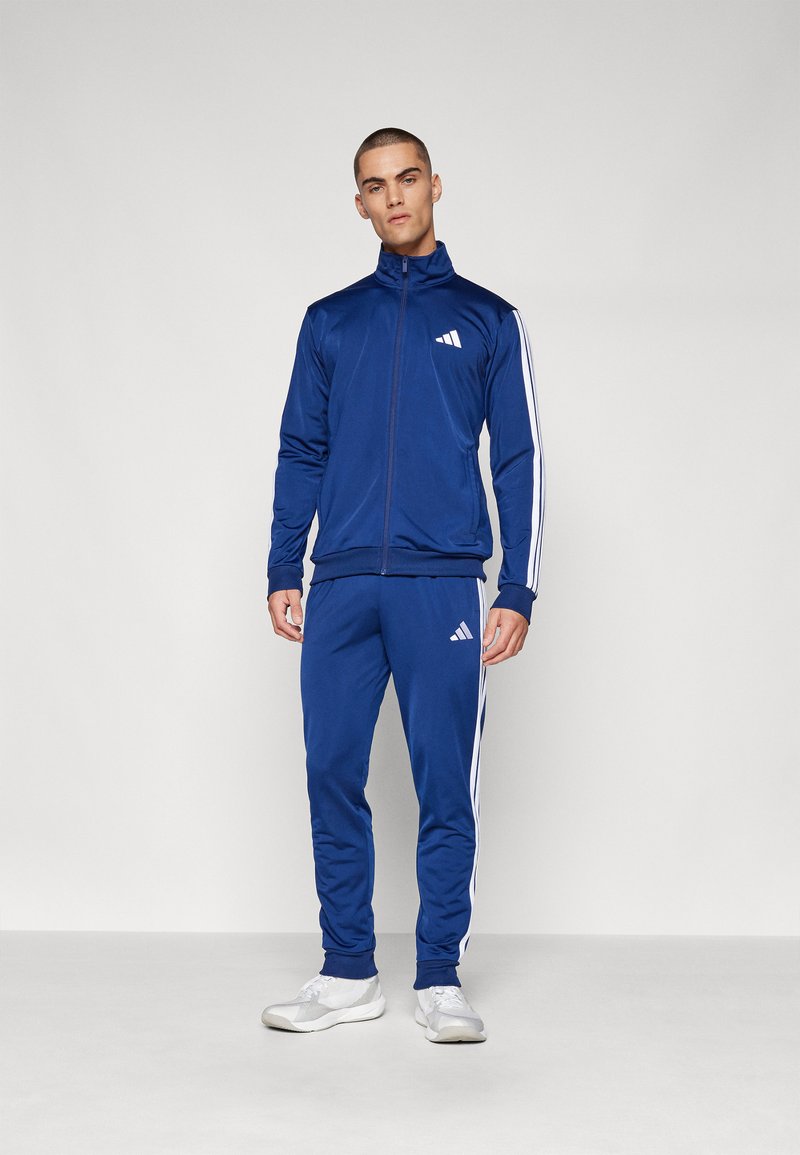 Modra trenirka iz gladkega materiala, ki ima bele črte ob straneh, z zip jopico in Adidasovo logo na prsih in hlačah.