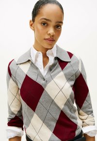 Maglione con motivo argyle grigio, borgogna e crema, con colletto e chiusura a tre bottoni, indossato sopra una camicia bianca. Texture morbida.