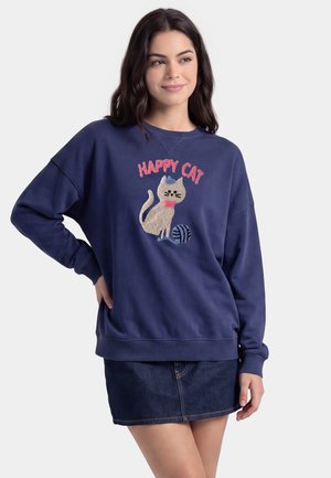 Giovane donna che indossa una felpa blu navy con un design "Happy Cat" e una gonna di jeans, in piedi con una mano sul fianco.