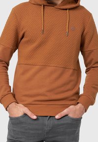 Mężczyzna z rękami w kieszeniach, ubrany w rust-colored bluzę z textured górną częścią oraz ciemnoszare spodnie, na jednolitym tle.