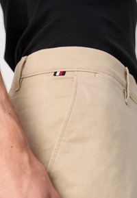 Pantaloni di cotone beige con fronte piatto, caratterizzati da una piccola etichetta logo rossa, bianca e blu sulla vita. Texture liscia, vestibilità sartoriale.