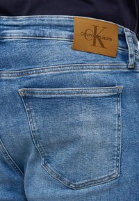 Jeans de denim azul desgastado con una etiqueta de cuero beige en la que está grabado "Calvin Klein Jeans", diseño de cinco bolsillos con detalles de costura.