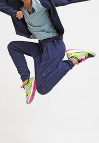 Marina träningsbyxor, ljusblå skjorta och färgglada sneakers med ett gult och rosa mönster. Figuren är i luften i ett hopp mot en enkel bakgrund.