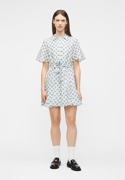 Femme debout portant une courte robe blanche à imprimé floral bleu, des chaussettes blanches et des mocassins noirs, sur un fond clair uni.