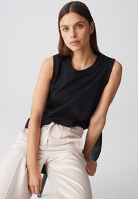 Woman with straight brown hair wearing a sleeveless black top and beige pants, sitting slightly angled on a chair against a plain background.