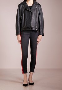 Kvinna som bär en svart läderbikerjacka över svarta tajta jeans med röda sidoremmar och svarta spetsiga klackskor, ståendes på ett beige golv.