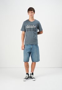 Grå t-skjorte med svart grafisk tekst, parret med lyse denimshorts og svarte høye sneakers med hvite lisser. Enkel og uformell antrekk.