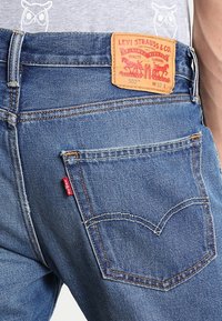 Blå denimjeans med en lapp märkt "Levi Strauss & Co." och en röd etikett. Synlig sömnad, klassisk femficksdesign och en blekt textur.