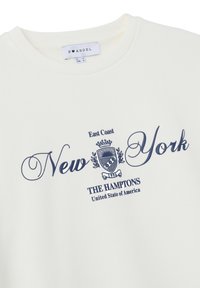 Sudadera blanca con texto en azul marino "Nueva York" y "Los Hamptons Estados Unidos de América" que presenta un escudo y "Costa Este" en la parte superior.