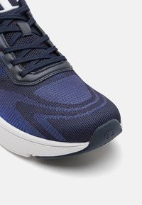 Marinblå sportskor med en texturerad tygövre, förstärkt tå och en vit mellansula med logodetalj på hälen.