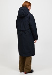Marineblauwe lange jas met een capuchon en een kortere achterkant, voorzien van een trekkoorddetail, gecombineerd met bruine broeken en zwarte schoenen.
