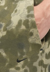 Zaļš kamuflāžas raksts audumam ar gludu tekstūru, kurā ir melns Nike logotips. Tuvplāns par materiālu un dizaina detaļām.