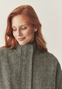 Cappotto grigio a quadri con colletto alto e tessuto texturizzato. Presenta un motivo sottile e linee pulite, enfatizzando una silhouette strutturata.