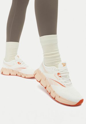 Białe buty sportowe z teksturowanymi siateczkowymi cholewkami, brzoskwiniowymi akcentami i masywnymi podeszwami z pomarańczowymi elementami; zestawione z kremowymi skarpetkami i brązowymi legginsami.