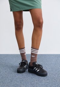 Svarta sneakers med tre vita ränder, matchade med bruna ribbade strumpor med vita ränder och logotypaccent. Grön kjol synlig ovanför.