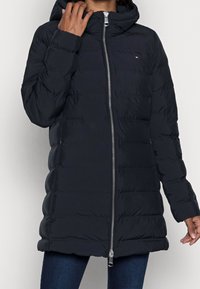 Tumma pusakka, jossa on vetskari edessä, huppu ja tikattu kuvio. Varustettu pienellä logolla ja sivutaskuilla. Valmistettu kevyestä materiaalista.