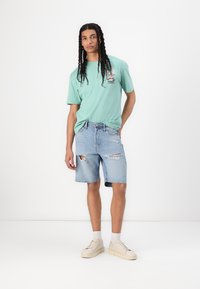 Tricou verde deschis cu imprimeu grafic, asortat cu pantaloni scurți din denim distresat și adidași bej. Modelul stă în fața unui fundal alb simplu.