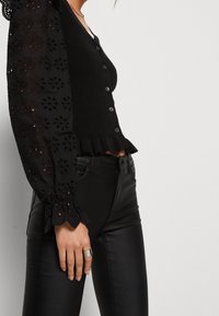 Svart ribbad cardigan med blommig ögla puffärm, knäppning fram och volangbotten; i kombination med svarta tajta byxor som har en silvrig ring.