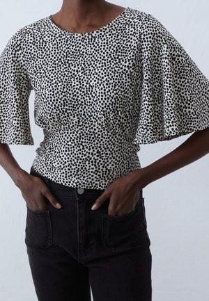 Zwarthuidig persoon draagt een witte blouse met zwarte onregelmatige stippen en brede fladdermouwen, gecombineerd met een zwarte broek met hoge taille, handen in de zakken.