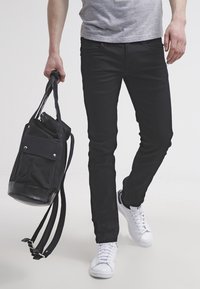 Černé slim-fit džíny doplněné o bílé tenisky. V jedné ruce drží plátěný batoh s koženým základem a přední kapsou.