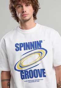 T-shirt en coton blanc avec un texte graphique audacieux en bleu et jaune qui dit "SPINNIN' GROOVE" avec des accents circulaires. Manches courtes, col rond.