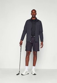 Čierna bunda na zips, čierne tričko a tmavé šortky. Nosenie bielych športových topánok, drží golfovú palicu. Jednoduchý dizajn a funkčné oblečenie.