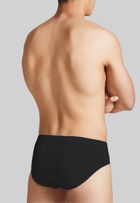Costumi da bagno neri con un design elegante, vita bassa, texture liscia e cuciture minime, visti da dietro su uno sfondo neutro.