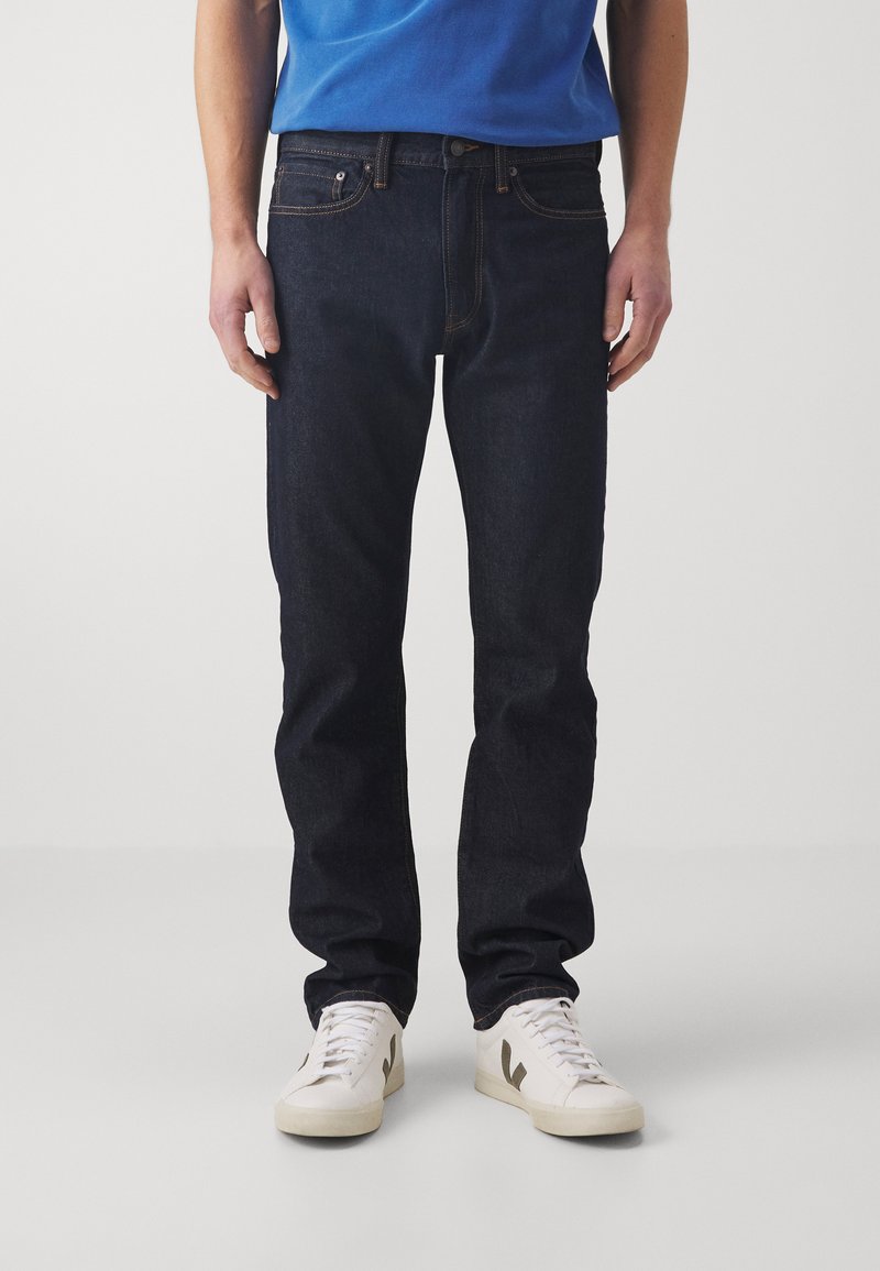 Pantalones vaqueros de mezclilla azul oscuro con corte recto, que cuentan con un estilo clásico de cinco bolsillos y costuras de contraste. Combinados con zapatillas blancas.