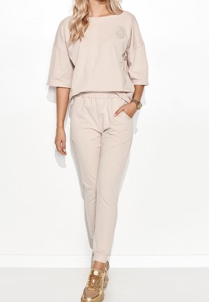 Kobieta ubrana w beżową, oversize'ową koszulkę z krótkim rękawem oraz pasujące dresowe spodnie, na stopach złote trampki, stoi z jedną ręką w kieszeni.