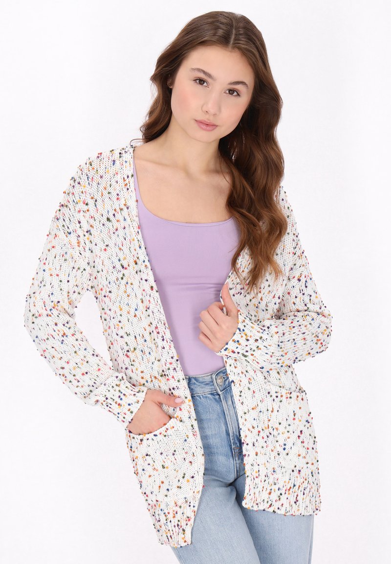 Gebreide cardigan met multikleurige spikkels op een witte achtergrond, open voorkant, lange mouwen en twee voorkruis zakken. Draagt over een paarse tanktop.