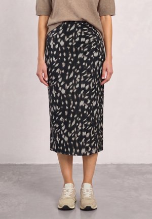 Femme portant une jupe midi noire et grise à motifs, un haut beige à manches courtes et des baskets beiges, debout sur un sol gris devant un mur uni.