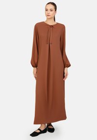 Bruine maxi-jurk met een ronde halslijn en strikdetail, met lange pofmouwen. Gemaakt van een soepele stof met een losse pasvorm.