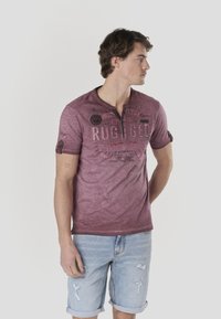 Chemise bordeaux à manches courtes avec col henley et motif graphique. Portée avec un short en denim clair déchiré.