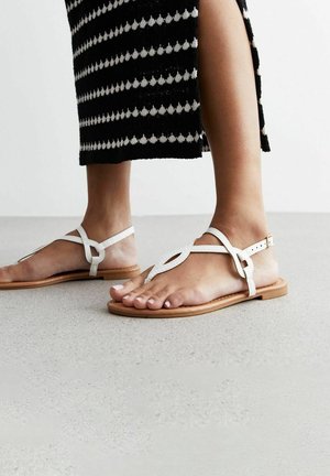 Pieds portant des sandales plates blanches à brides avec boucles à la cheville, associées à une jupe à motifs noir et blanc avec une fente sur le côté.