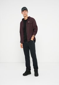 Camicia di flanella a quadri bordeaux sopra una maglietta nera, abbinata a jeans neri e sneaker nere. Il modello indossa un cappellino nero.