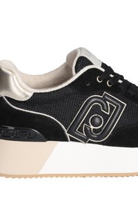 Baskets noires avec une tige en mesh et des accents en daim, un talon métallique et un logo distinct sur le côté. Semelle épaisse blanche avec un motif texturé.