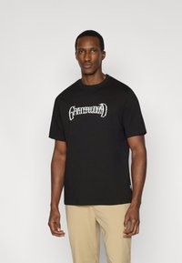 Μαύρο βαμβακερό T-shirt με λευκό γραφικό σχέδιο, στρογγυλή λαιμόκοψη, κοντά μανίκια και ίσια ραφή. Φορεμένο με ταμπά παντελόνια.