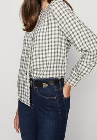 Blouse à carreaux noir et blanc avec des manches longues, devant boutonné, associée à un jean en denim bleu foncé et une ceinture noire avec des accessoires en métal.