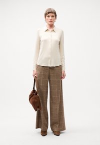 Beige Hemd mit Knöpfen, braune karierte weite Hose, braune Wildlederstiefel mit Absatz und eine braune Stoffhandtasche mit Zugverschluss.