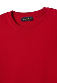T-shirt rossa con colletto girocollo, dotata di un'etichetta nera visibile con la scritta gialla "PIOMBO" all'interno del collare.