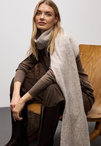 Pull en tricot marron avec des manches côtelées, associé à une écharpe texturée beige clair. Le mannequin est assis sur une chaise en bois portant des bottes en cuir noir.