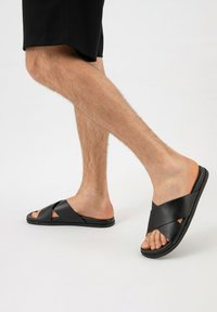 Sandales glissantes en cuir noir avec des brides croisées, semelle plate, design à bout ouvert et texture lisse. Portées avec un short noir.