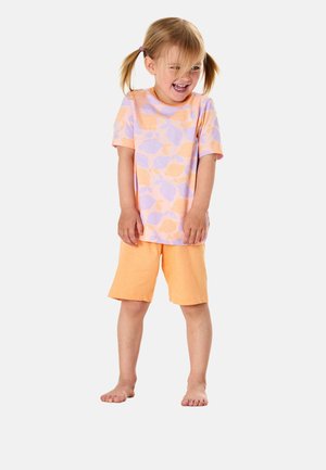 Young girl with blonde pigtails laughing, wearing a purple and orange patterned t-shirt and orange shorts, standing barefoot on white background.