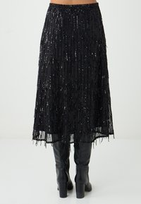 Gonna nera con frange e paillettes, caratterizzata da un design alla lunghezza del ginocchio e una finitura testurizzata. Abbinata a stivali neri fino al ginocchio.