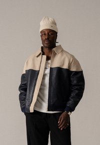 Læderbomberjakke med en beige overdel og navy nederste sektion, med lynlåsfortagning, sidelommer og ribbede manchetter. Modellen bærer en strikket hat.